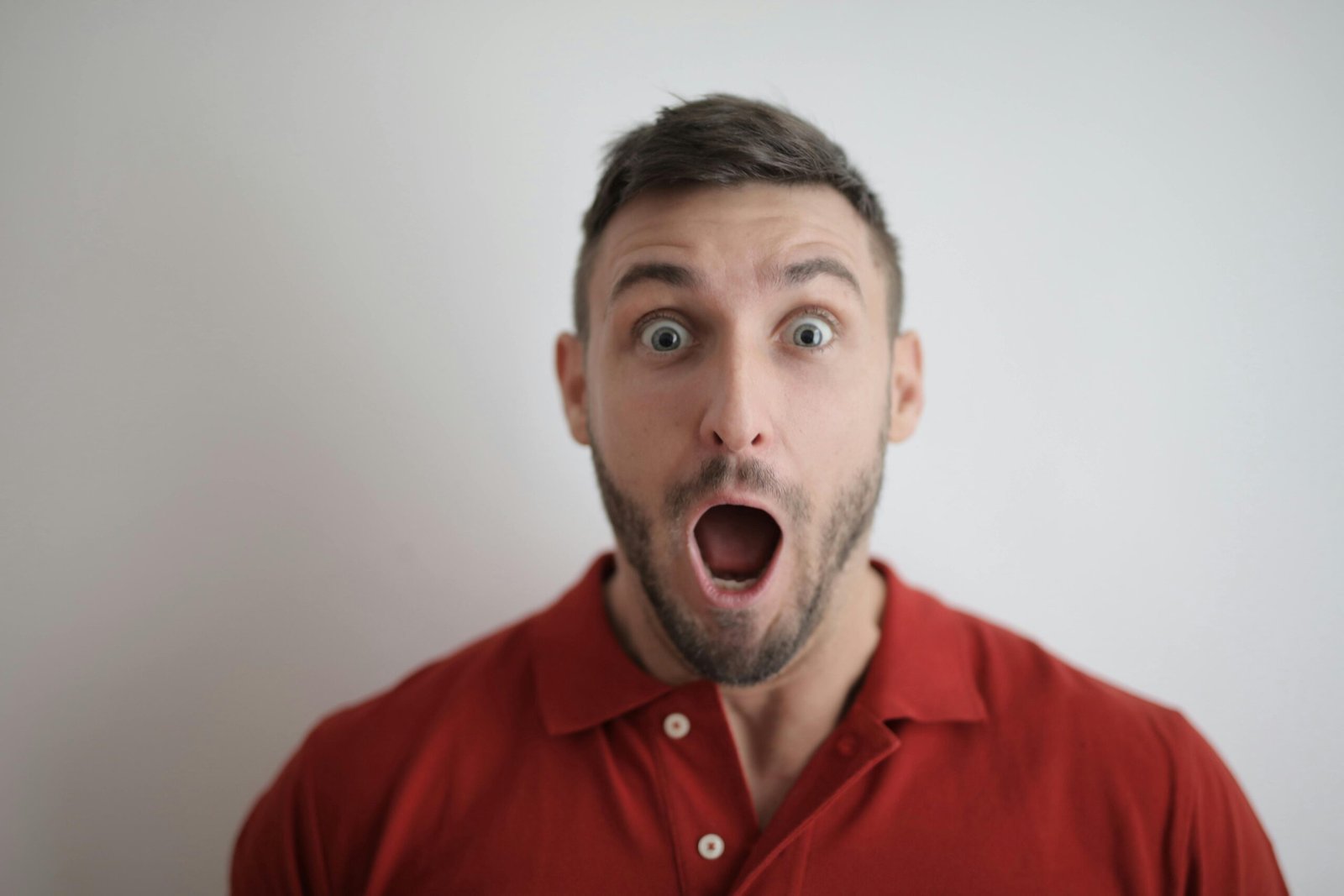 Portrait of a shocked man in a red polo shirt with a white background, expressing surprise.